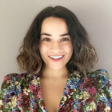 A person with shoulder-length wavy hair smiles at the camera. They are wearing a colorful floral top with a neutral background behind them.