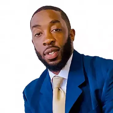 A man with a beard wearing a blue suit and a light-colored tie against a plain white background. He is looking toward the camera.