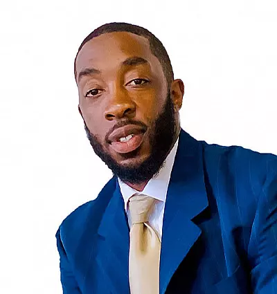A man with a beard wearing a blue suit and a light-colored tie against a plain white background. He is looking toward the camera.