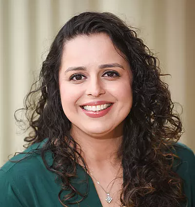 A woman with long, curly dark hair smiles warmly at the camera. She is wearing a dark green top and a delicate necklace with a pendant. The background is softly blurred with neutral tones.