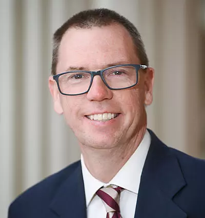 A smiling man with short hair, wearing glasses and a formal suit with a striped tie. The background is softly blurred, emphasizing his professional appearance.