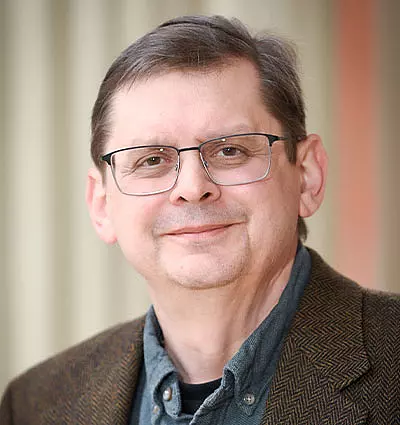 A middle-aged man with glasses smiles slightly. He is wearing a brown herringbone jacket over a gray shirt. The background is softly blurred with beige and orange tones.