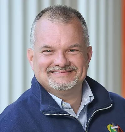 A man with short hair and a beard smiles at the camera. He is wearing a navy blue jacket over a light-colored shirt. The background features vertical white and orange stripes.