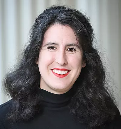 A person with long, wavy black hair and red lipstick is smiling at the camera. They are wearing a black turtleneck sweater. The background is softly blurred with vertical lines.