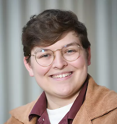 A person with short hair and glasses smiles warmly. They are wearing a brown jacket over a burgundy shirt. The background is softly blurred with vertical lines.