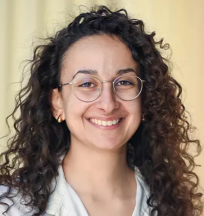 Smiling person with curly hair, wearing glasses and a white shirt, stands in front of a neutral background.