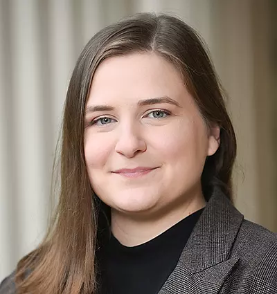 A person with long brown hair is smiling softly, wearing a dark top and a gray blazer. The background is softly blurred with vertical lines.
