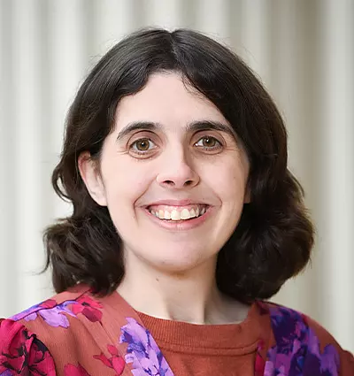 A person with shoulder-length dark hair smiling, wearing a floral-patterned blouse. The background features vertical light-colored panels.