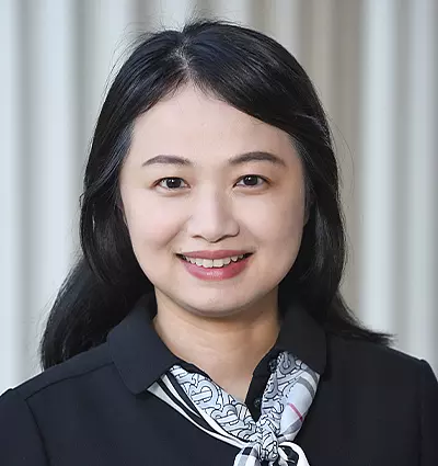 A smiling woman with long dark hair wearing a black top and a patterned scarf, standing against a light-colored background featuring vertical lines.