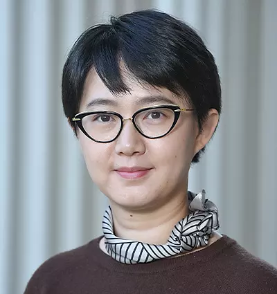 A person with short black hair and glasses smiles softly at the camera. They are wearing a dark top and a black-and-white patterned scarf around their neck, standing against a neutral, blurred background.