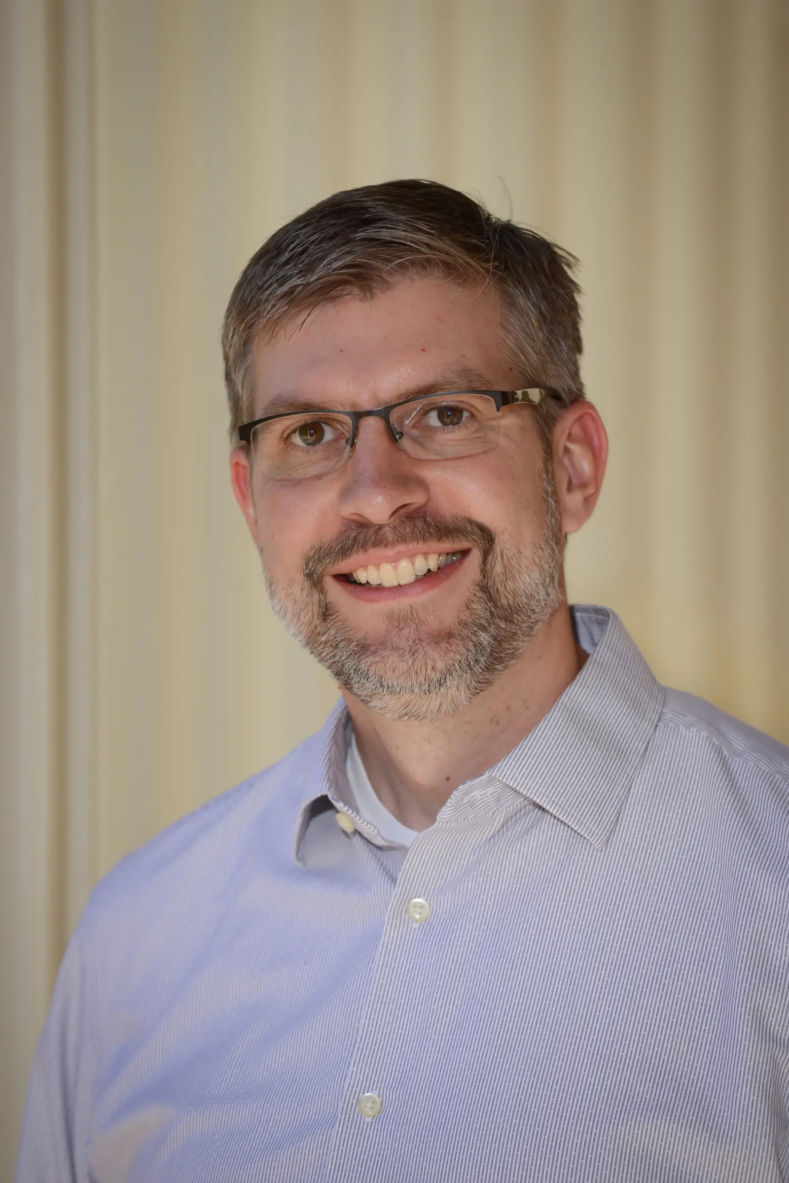 A man with short hair and glasses smiles at the camera, wearing a light striped button-up shirt. The background is softly blurred, creating a neutral and warm tone.