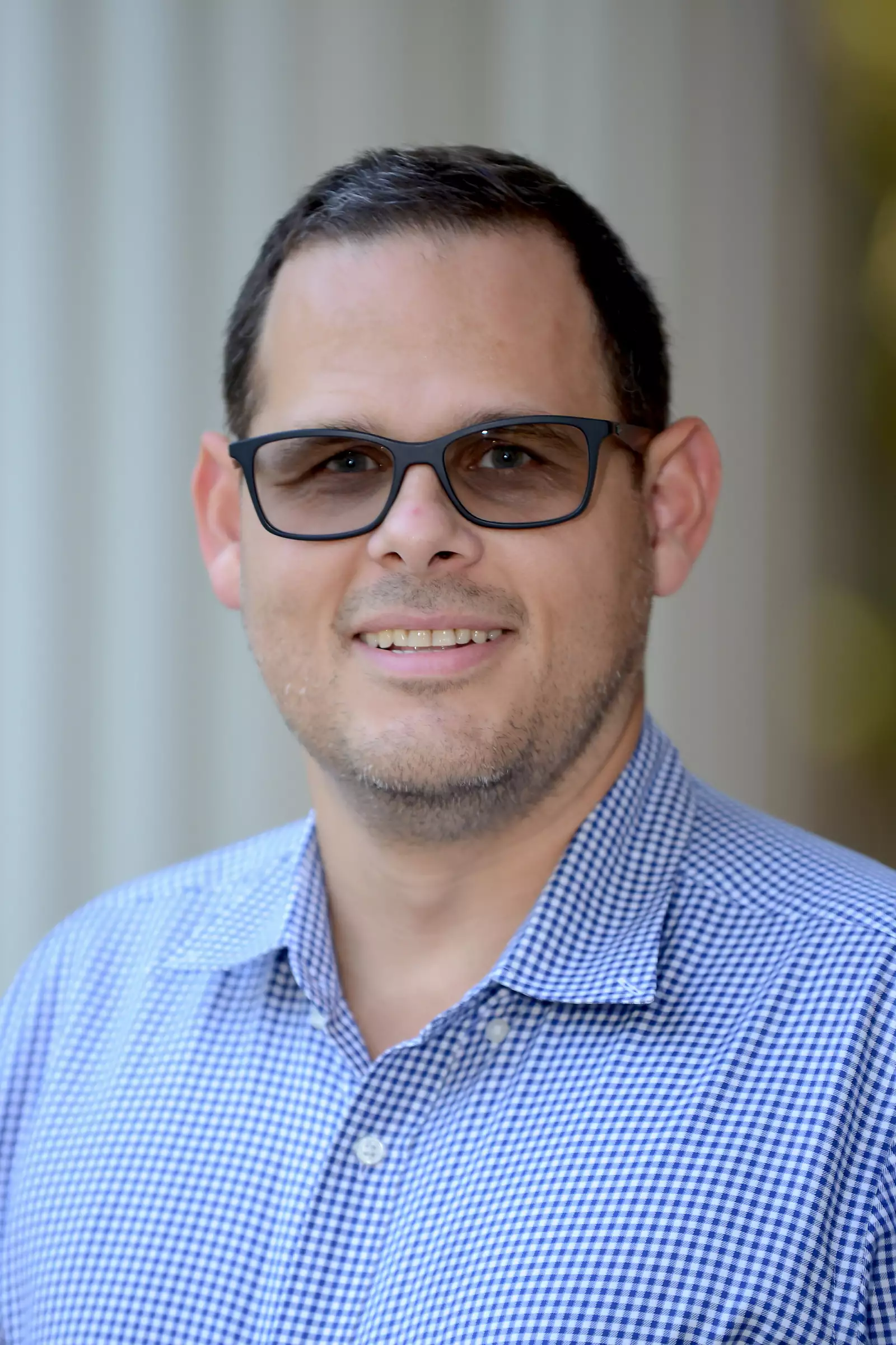 A man wearing black glasses smiles at the camera. He has short dark hair and is dressed in a blue and white checkered shirt. The background is softly blurred, with vertical lines suggesting pillars or columns.