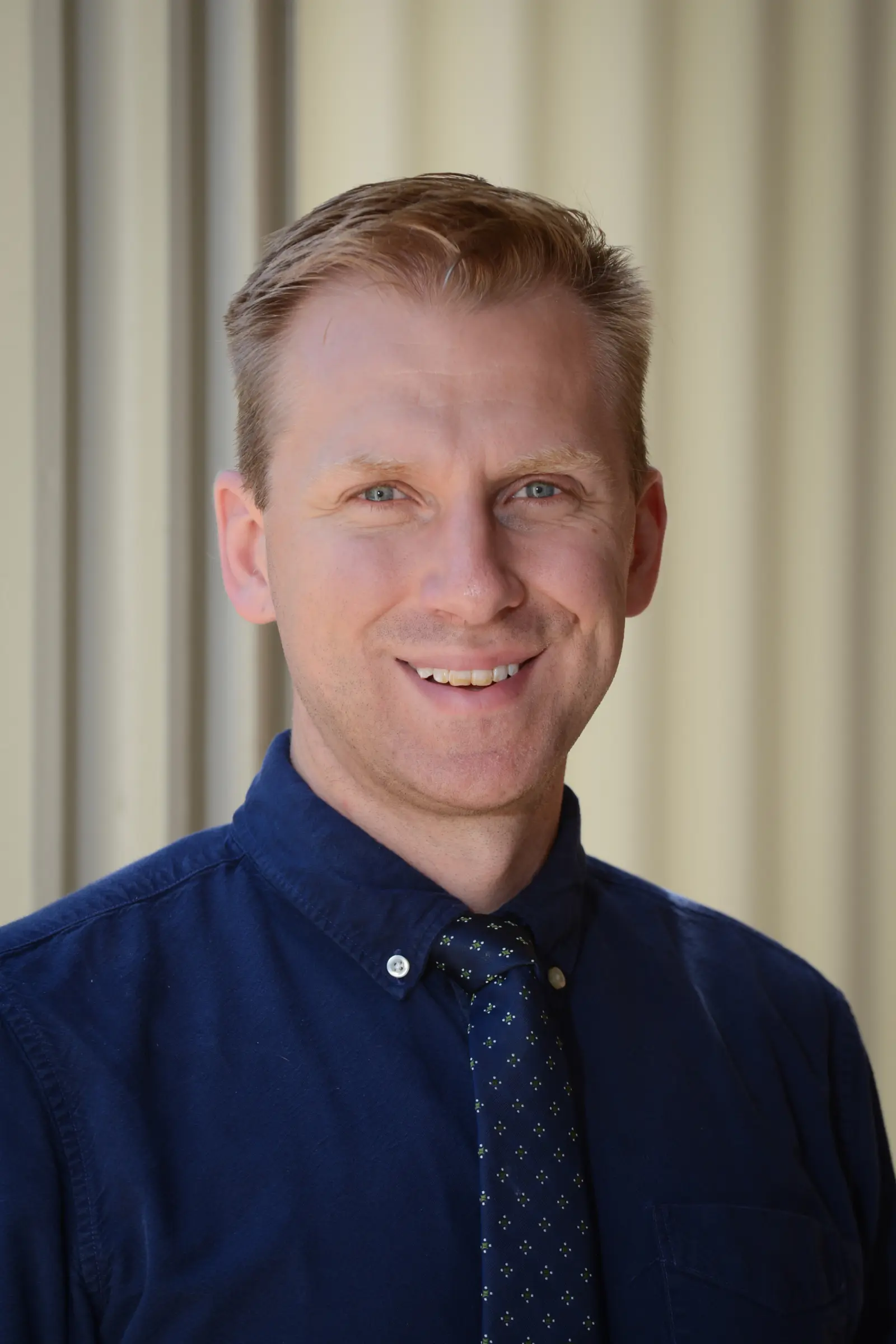 A smiling person with short, light-colored hair, wearing a dark blue button-up shirt and a tie with small polka dots. The background is softly blurred, showing vertical lines.