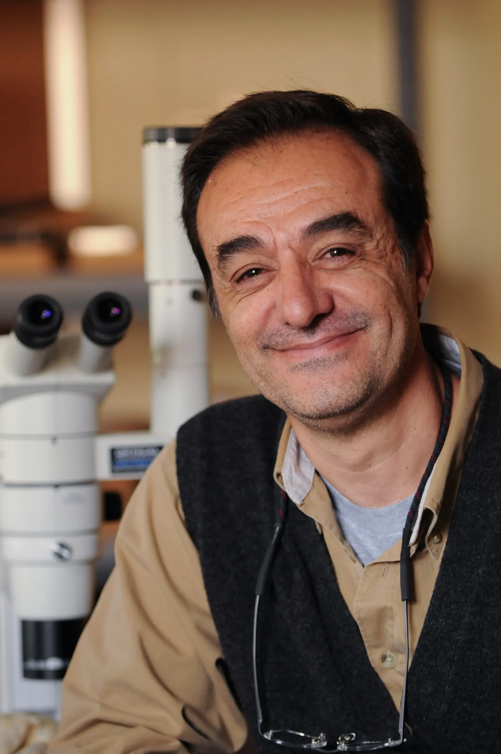 A person smiling at the camera, sitting next to a microscope in a laboratory setting. They are wearing a dark vest over a beige shirt and have reading glasses hanging from their neck.