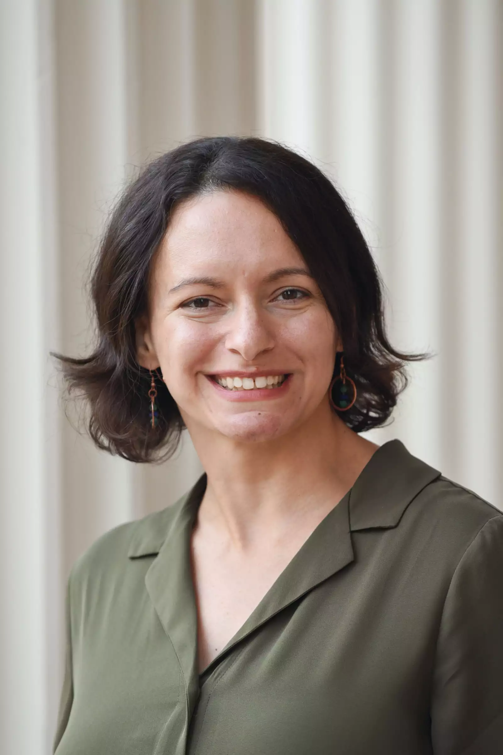 A person with short, dark hair and earrings smiling, wearing an olive green top, stands in front of a light-colored, vertically striped background.