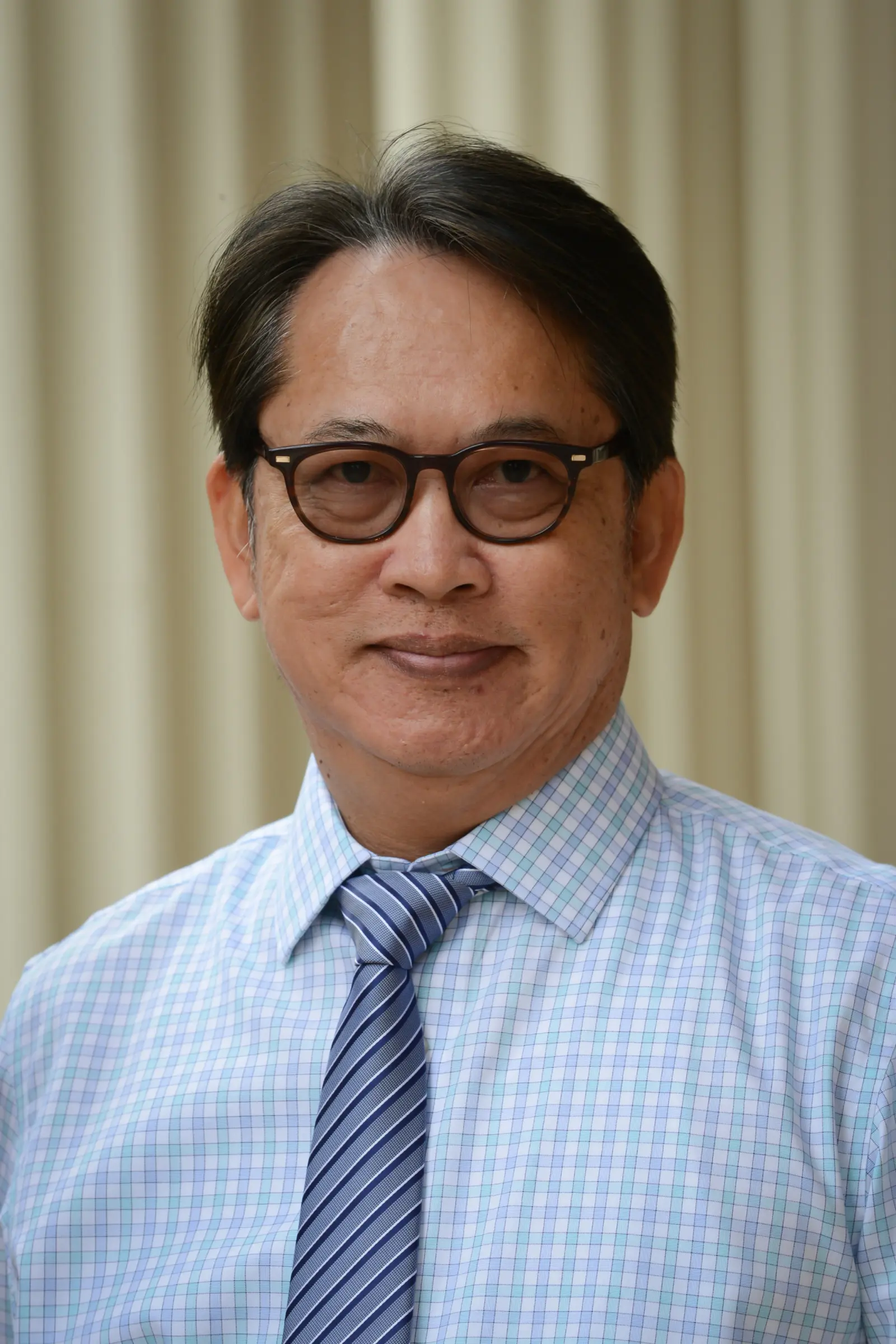 A person with short hair and glasses is wearing a blue and white checkered shirt with a striped tie. The background is blurred beige vertical lines.