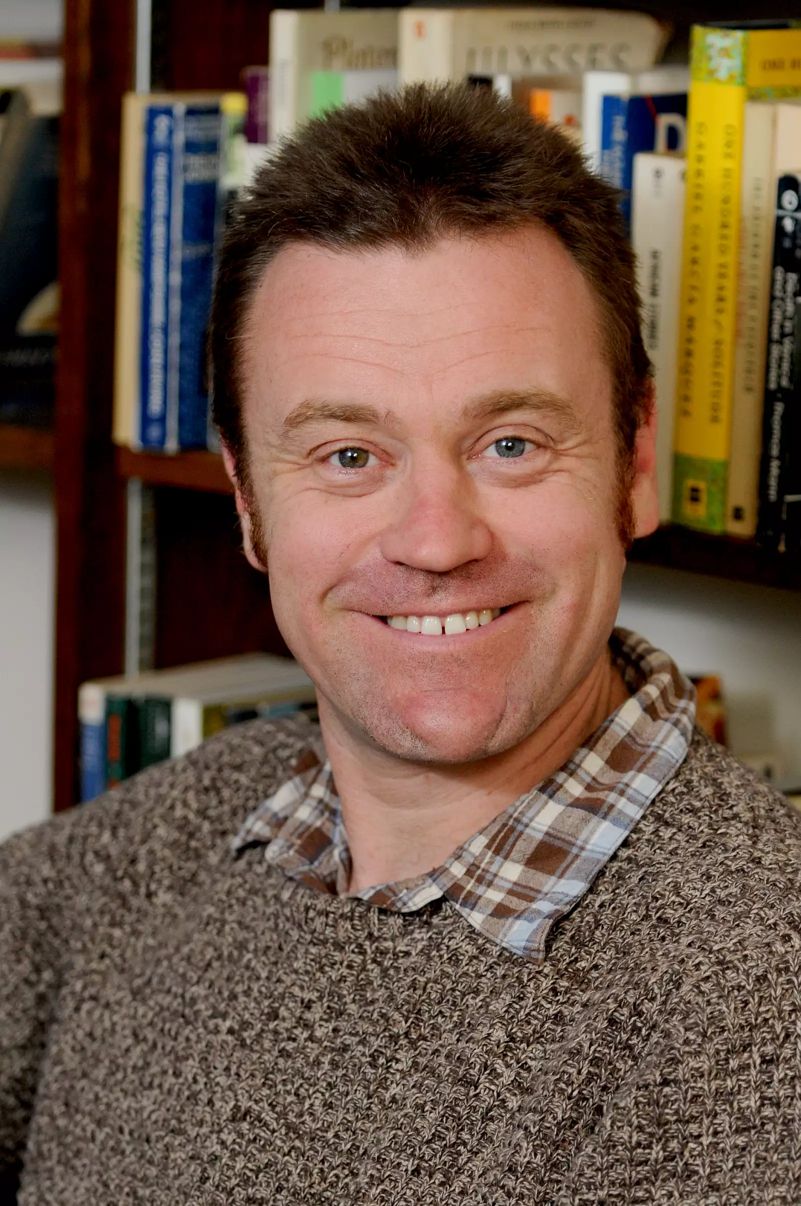 A smiling man with short dark hair, wearing a plaid shirt under a brown sweater, is sitting in front of a filled bookshelf.