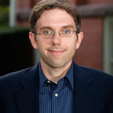 A man with short brown hair and glasses in a dark suit and striped shirt stands in front of a brick building, smiling at the camera.
