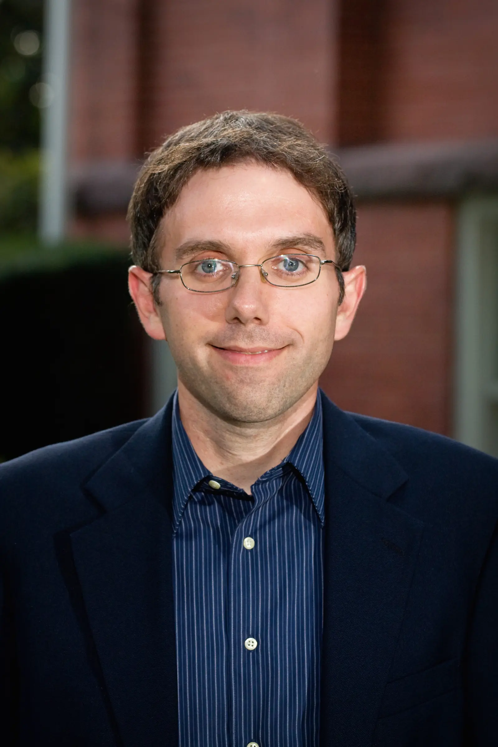 A man with short brown hair and glasses in a dark suit and striped shirt stands in front of a brick building, smiling at the camera.