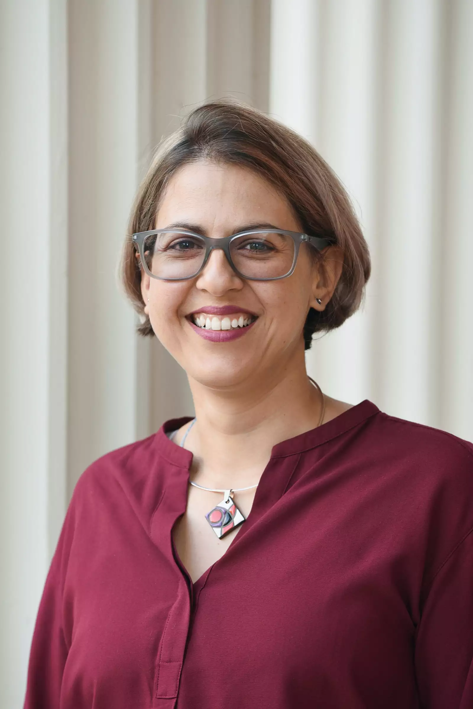 A person with short hair and glasses is smiling, wearing a maroon blouse and a geometric necklace, standing in front of a light-colored, vertically striped wall.