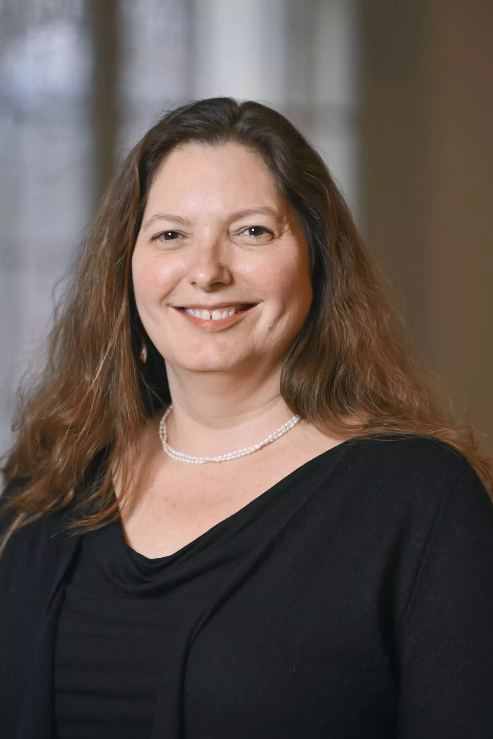 A smiling woman with long brown hair wearing a black top and a pearl necklace stands in front of a blurred background.