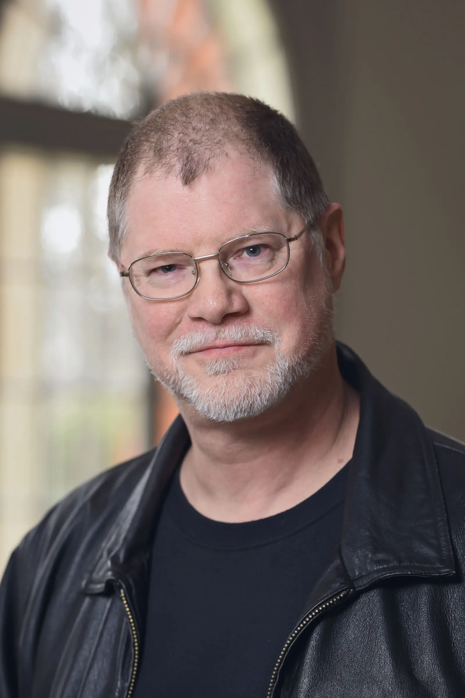 A man with short hair and glasses, wearing a black leather jacket over a black shirt, smiles softly. He has a trimmed beard and stands in front of a window with a soft, blurred background.