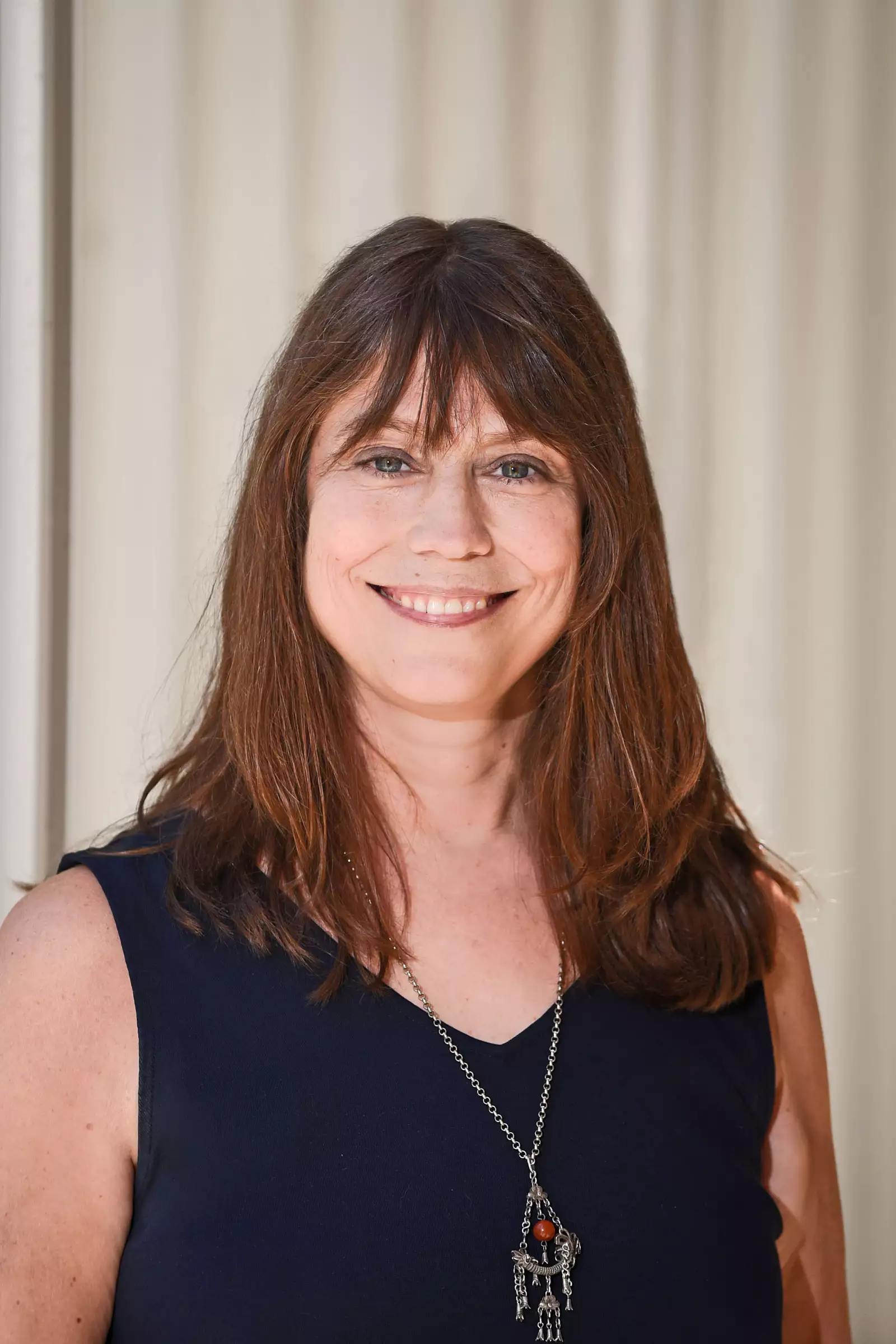 A woman with long brown hair smiles at the camera. She is wearing a sleeveless navy blue top and a necklace with a pendant. The background is a light-colored curtain.