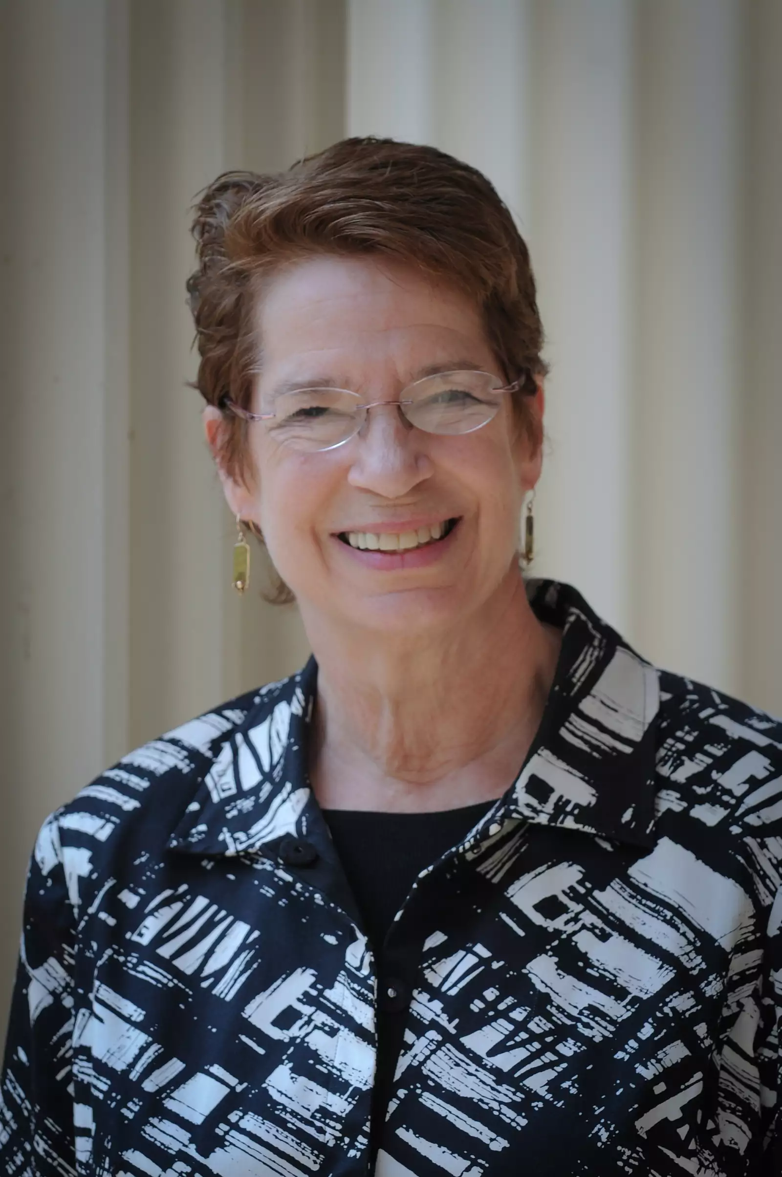 A smiling woman with short, brown hair wearing glasses and a patterned black and white jacket stands in front of a neutral background.