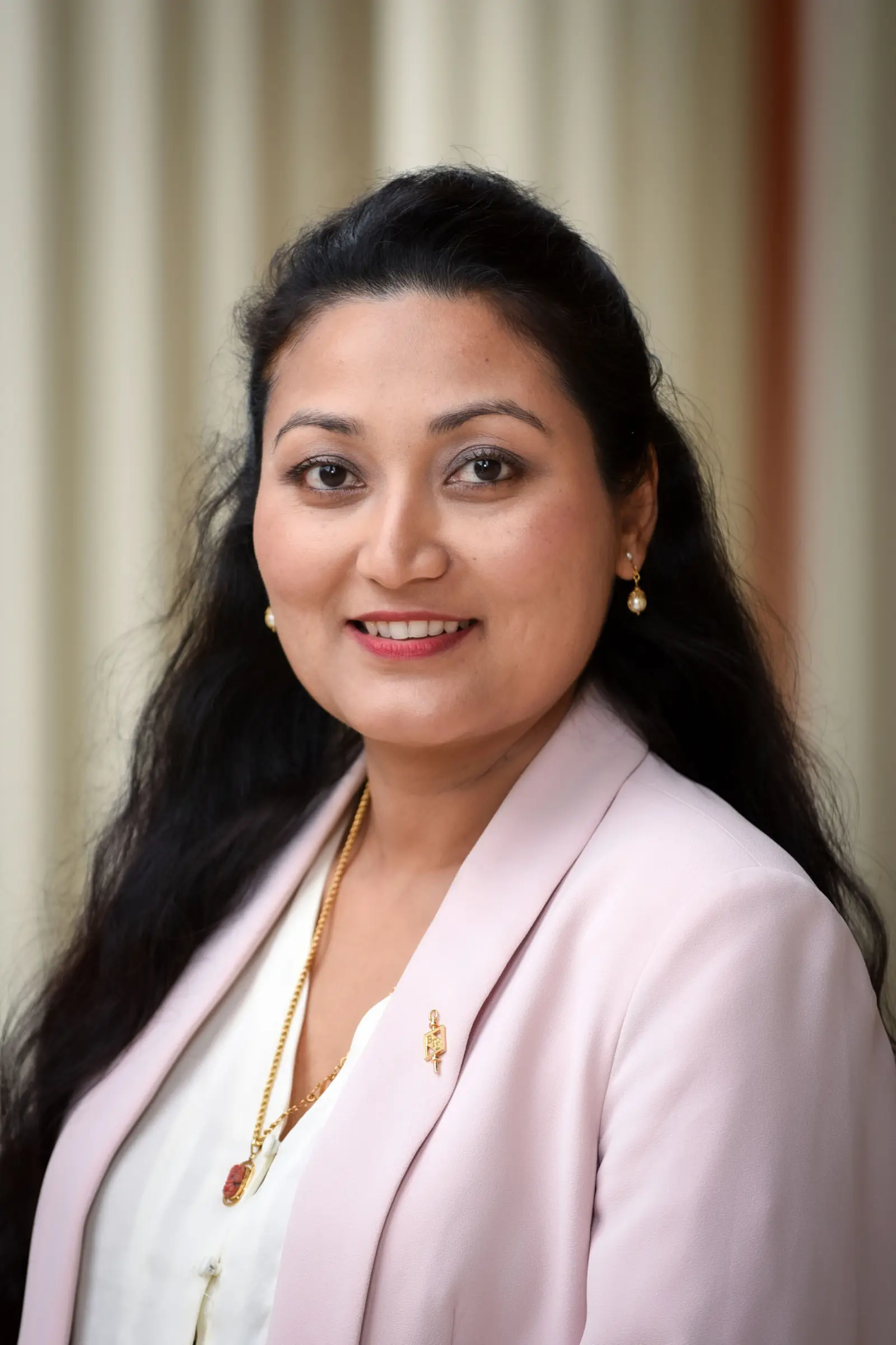 A woman with long, dark hair is smiling. She is wearing a light pink blazer over a white blouse, accessorized with gold earrings and a necklace. The background features light-colored vertical stripes.