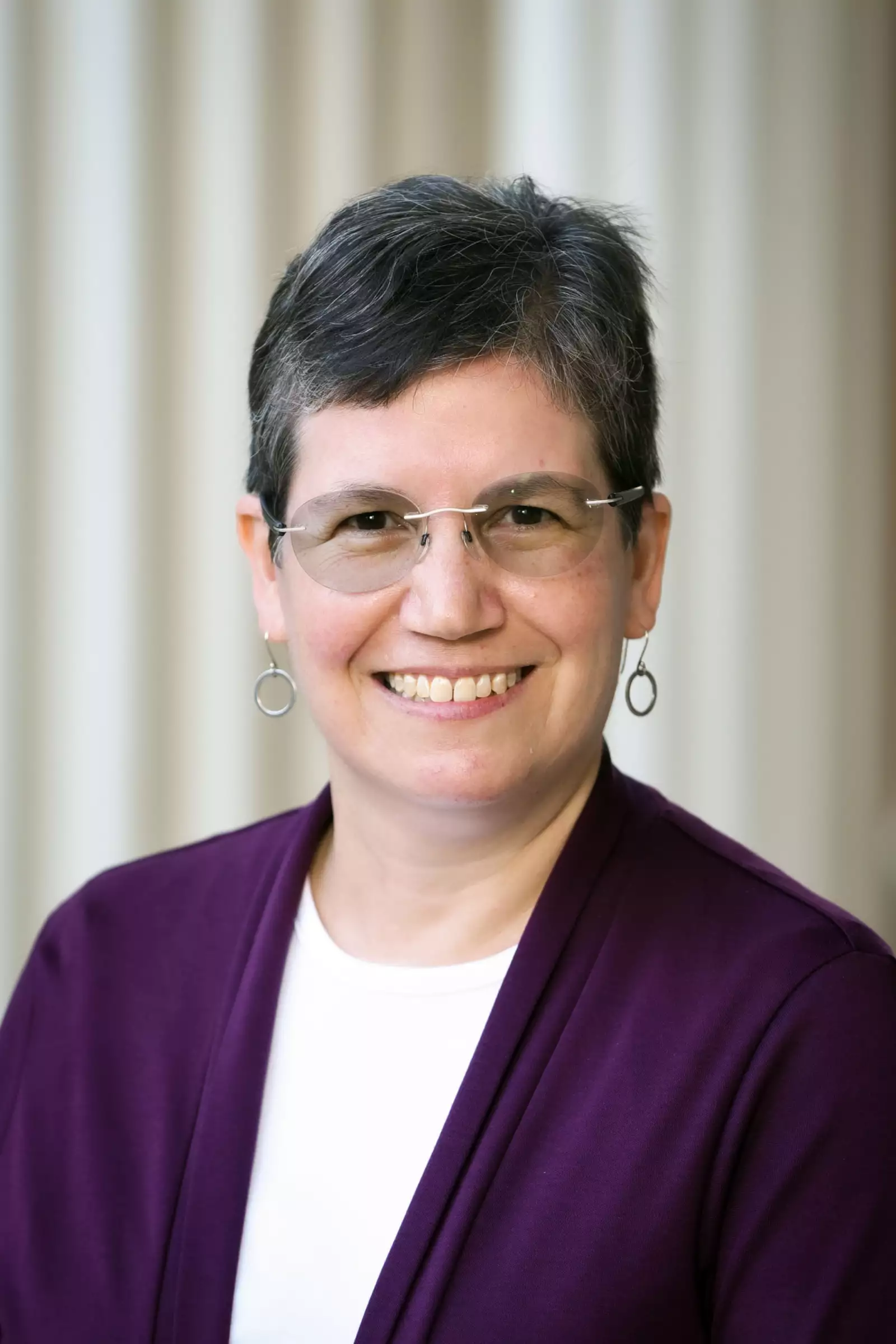 A person with short gray hair and glasses smiles at the camera. They are wearing a purple cardigan over a white shirt. The background features beige vertical stripes.