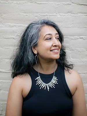 A woman with long dark and gray hair smiles while looking to the side. She is wearing a black sleeveless top, geometric earrings, and a statement necklace with vertical metal bars. The background is a light-colored brick wall.