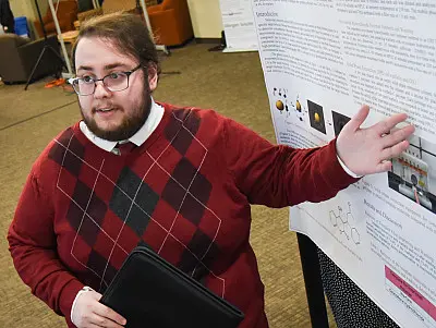 A person with glasses, wearing a red argyle sweater and tie, is presenting research on a poster in an indoor setting. They are holding a folder and gesturing towards the poster, which contains text and images.