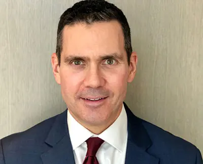 Man in a suit and tie with short dark hair, smiling slightly, standing against a neutral, textured background.