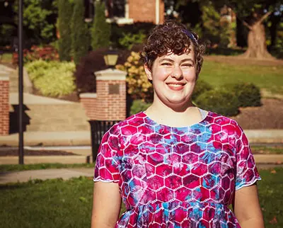 A person with short curly hair, wearing a colorful hexagon-patterned dress, smiles while standing outdoors. The background includes greenery, a brick structure, and a lamp post.