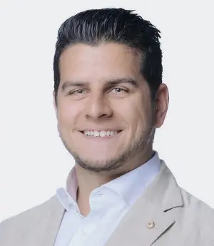 A man with short dark hair and a beard is smiling. He is wearing a light-colored suit jacket over a white shirt. The background is plain and light gray.