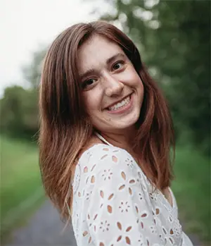A person with long brown hair smiles at the camera. They are wearing a white, patterned top. The background is a blurred outdoor setting with greenery.