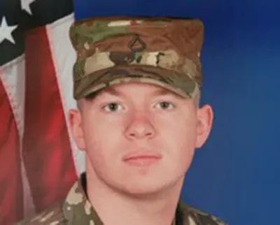 A person in military uniform with a camouflage cap stands in front of a backdrop featuring the American flag. The individual is looking directly at the camera with a neutral expression.