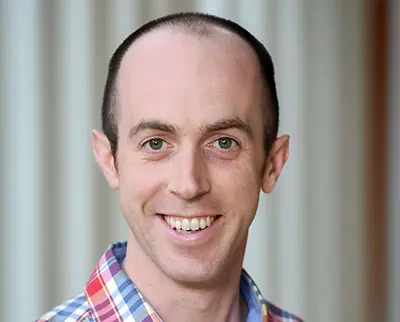A smiling person with short hair and a checkered shirt stands in front of a blurred background.