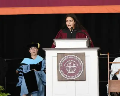 A person in a maroon robe speaks at a podium with a university emblem, during a graduation ceremony. Another person in academic regalia sits nearby.