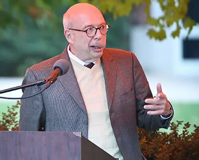 An elderly man in glasses and a suit speaks into a microphone at a podium outdoors. He gestures with one hand, and there is greenery in the background.