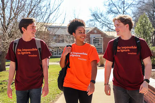 Three students walk outdoors wearing t-shirts with the slogan "Be impossible to ignore." Two have maroon shirts, and one has an orange shirt. They are smiling and carrying backpacks, with trees and a building in the background.