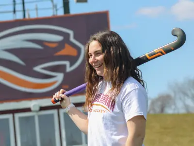 A smiling person stands on a field, holding a field hockey stick over their shoulder. They are wearing a white shirt with text. A large sign with an abstract eagle logo is visible in the background.