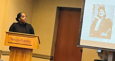 A person stands at a podium labeled "Susquehanna University" giving a presentation. A projected image of a historical figure in black and white is visible on a screen to their right.