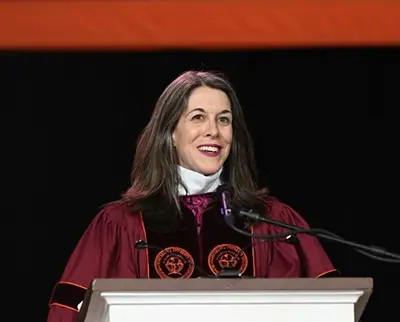 A person with long dark hair, wearing academic regalia and a white collar, stands smiling at a podium with microphones against a dark background. Orange and black drapes are visible above.
