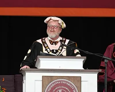 A person in academic regalia, including a cap and gown, stands at a podium with a microphone, delivering a speech. The podium features a logo for Susquehanna University, and a maroon and orange backdrop is visible.