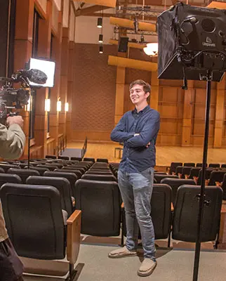 A person stands confidently in a lecture hall, with arms crossed, smiling at a camera crew. The room is equipped with stage lighting and rows of empty seats.