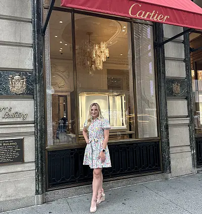 A woman in a floral dress stands smiling in front of a Cartier store with a red awning. The background features a decorative building facade and a large window displaying chandeliers inside.