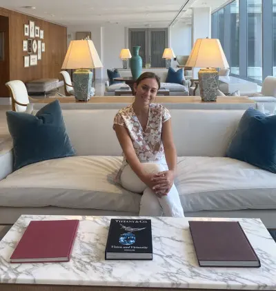 A person sits on a white sofa in a modern, well-lit living room with large windows. The room features decorative lamps and cushions. A marble coffee table in front holds three books, including one titled "Tiffany & Co.