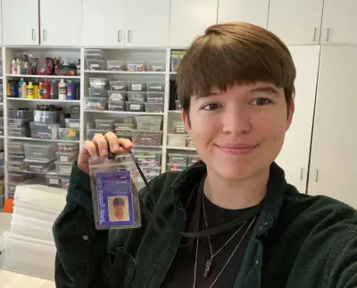 A person with short hair and a green shirt smiles at the camera while holding up an ID badge. Behind them are shelves filled with various colorful containers and supplies.