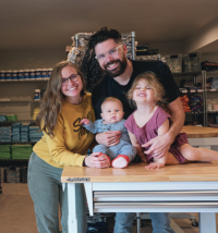 A smiling family of four gathers around a wooden table. The parents, wearing glasses, hold a baby and a young child. Shelves filled with various items are visible in the background, giving a cozy, homey atmosphere.